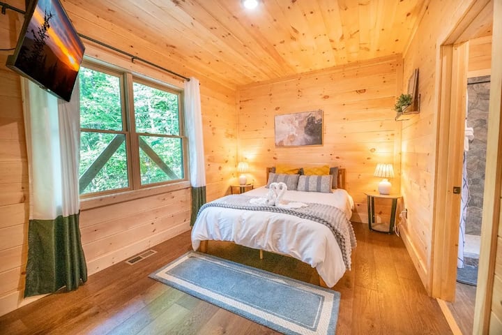 Relax in this tranquil bedroom featuring a comfortable bed, attached full bathroom and beautiful wood paneling. Enjoy the bright views from the large window and convenient entertainment with the mounted Smart TV.