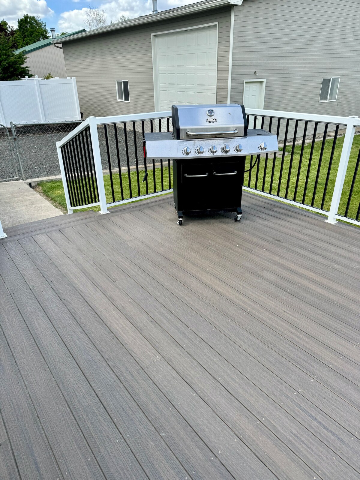 A spacious deck is presented, featuring a black gas grill positioned on a smooth surface. The deck railing is white, providing contrast against the grey backdrop of the nearby building and fenced yard. Green grass extends beyond the deck area.