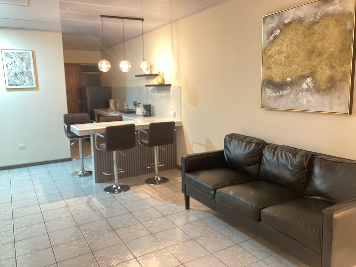 A modern kitchen and living area are shown with sleek design elements. A dark leather couch is positioned nearby, while the kitchen features a bar with high stools. Soft lighting from three pendant lights creates a welcoming ambiance, and light reflects off the tile flooring.