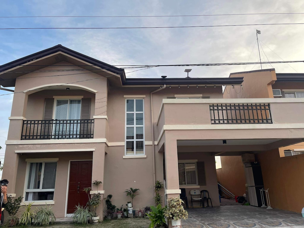 The two-story home features a balanced facade with light beige walls and dark brown accents. A covered balcony with a railing offers additional outdoor space. An entrance door is flanked by windows, while a paved area provides parking beneath the building.