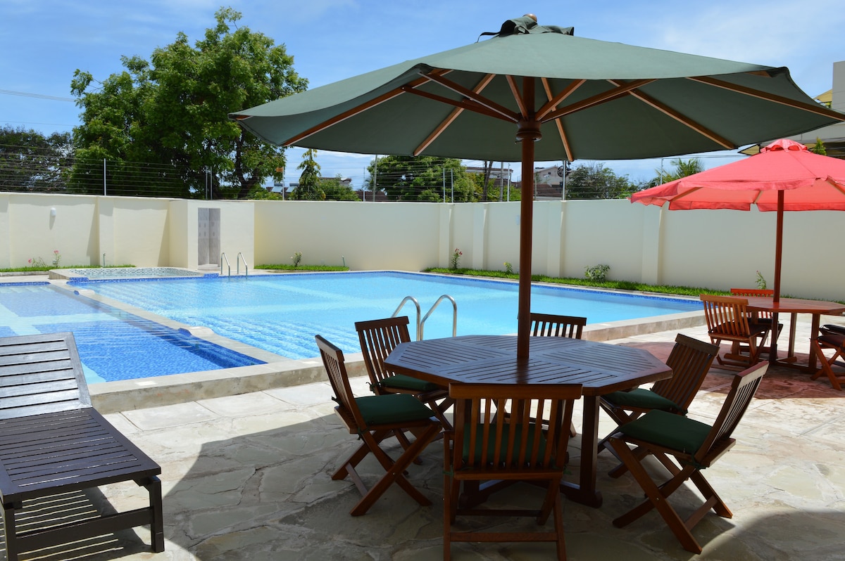 An outdoor pool area features a large swimming pool surrounded by a stone patio. Wooden tables with chairs are situated under green and red umbrellas, providing shaded seating. Lush greenery and a fence can be seen in the background.