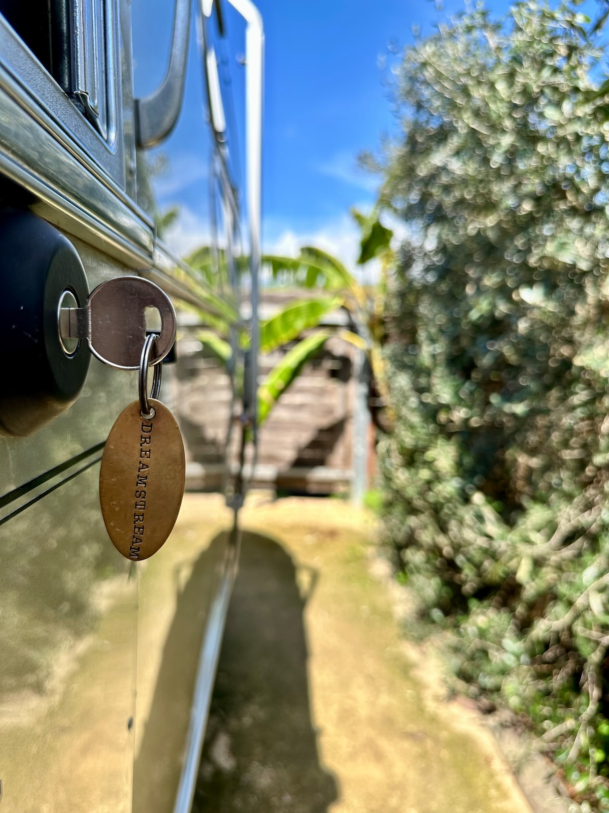 The image captures a close-up of a metal door handle with a key attached, featuring a wooden tag inscribed with 'Dreamer Airstream.' Sunlight filters through a lush green setting, highlighting the textured foliage and the reflective surface of the Airstream.