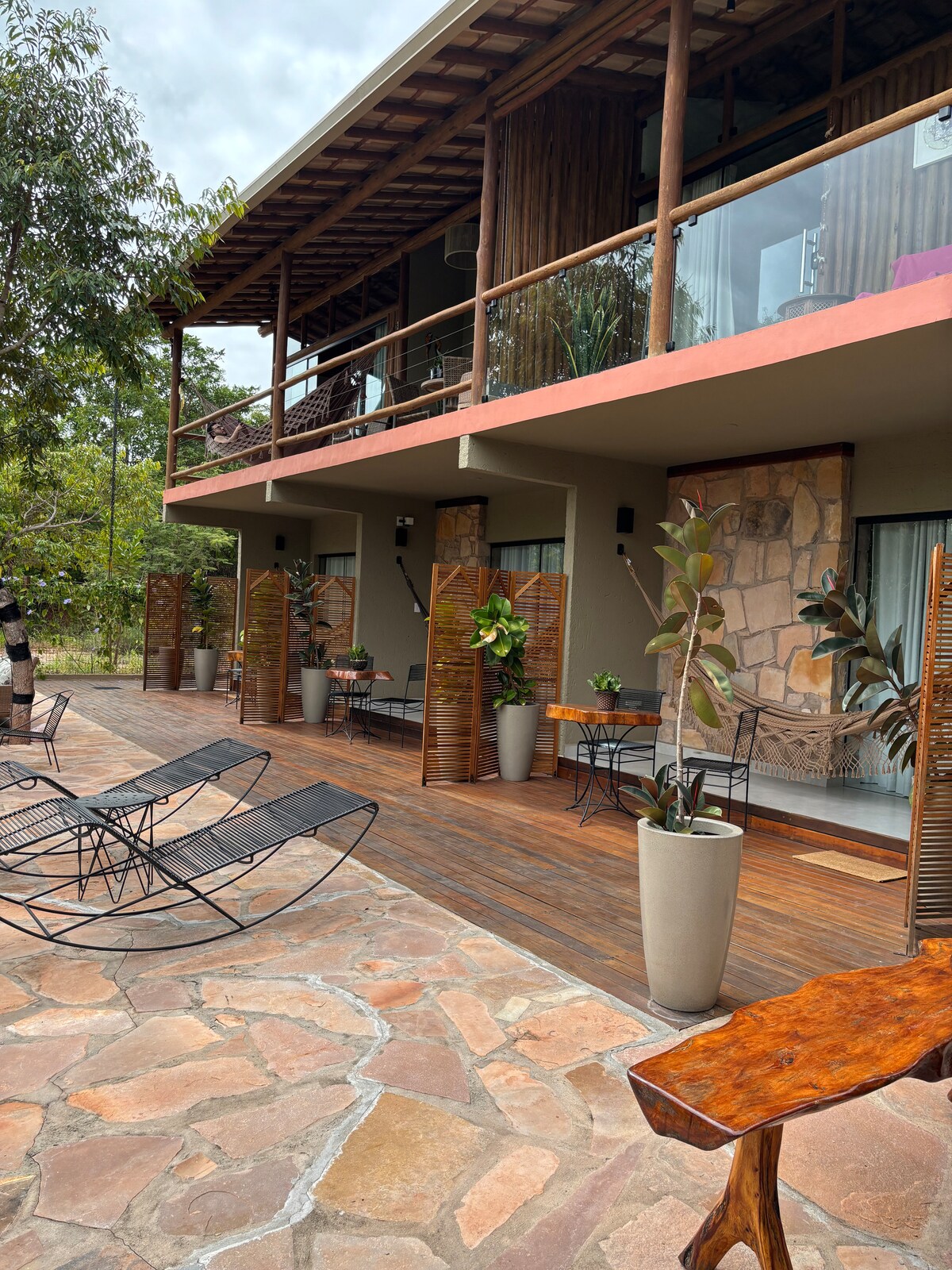 An inviting outdoor area features wooden decking surrounded by natural stone. Elegant lounge chairs are positioned to face the lush surroundings, while planters with green foliage add a touch of nature. The structure above includes large glass doors that open onto the deck.