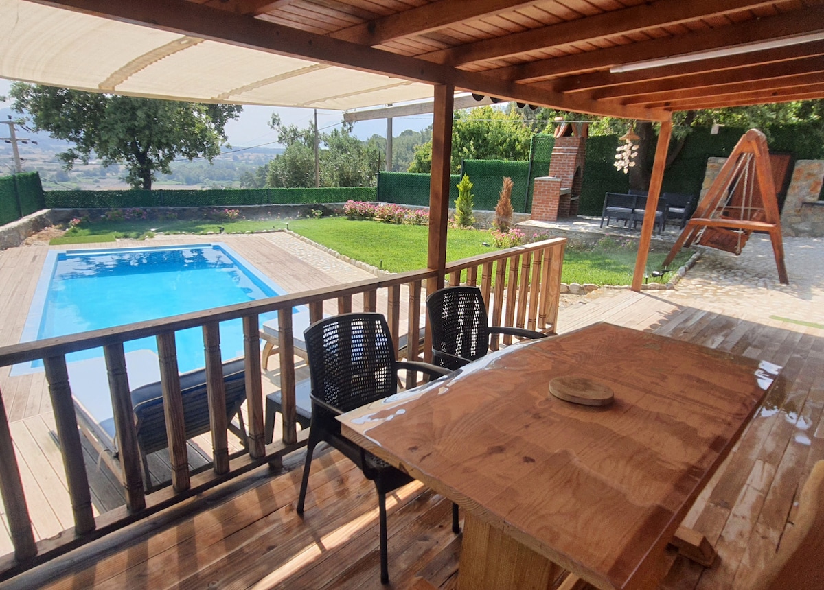 A wooden terrace showcases a table and two black chairs, overlooking a private pool surrounded by greenery. The scene includes a play area with a swing, with a fence in the background providing privacy. Sunlight highlights the inviting outdoor space.
