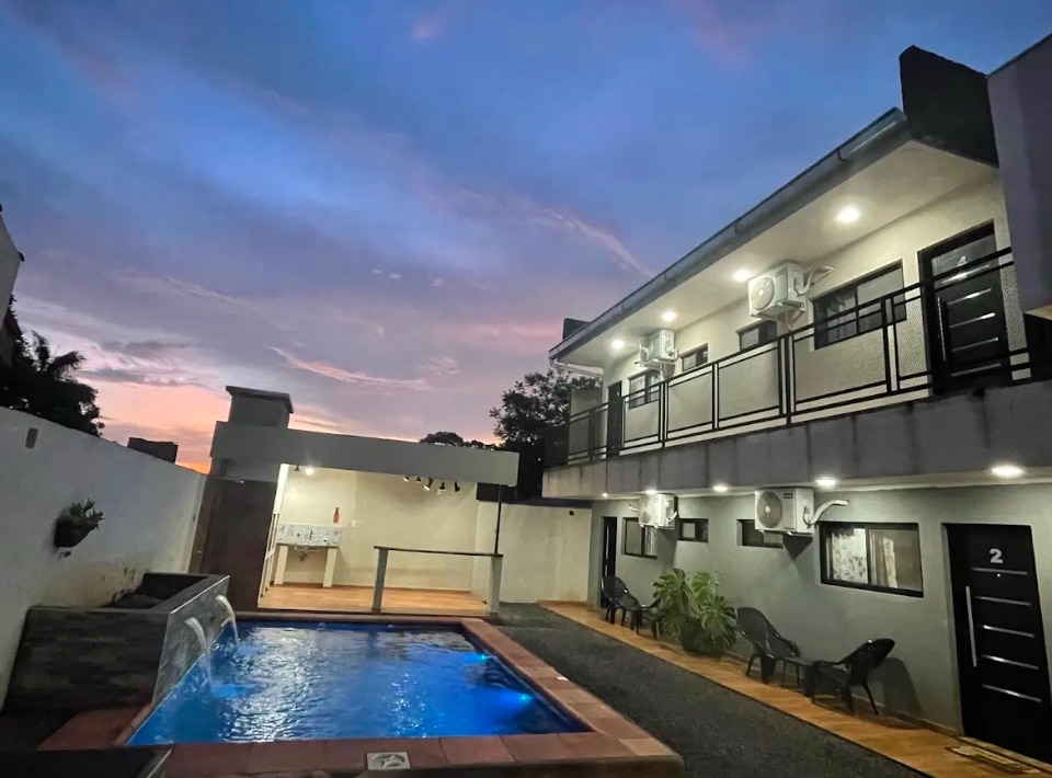 An inviting outdoor space features a swimming pool surrounded by wooden decking, with lounge chairs positioned nearby. The two-story building showcases balconies with black railings, and a covered outdoor area is visible, equipped with amenities for gatherings. The sky transitions to twilight hues.