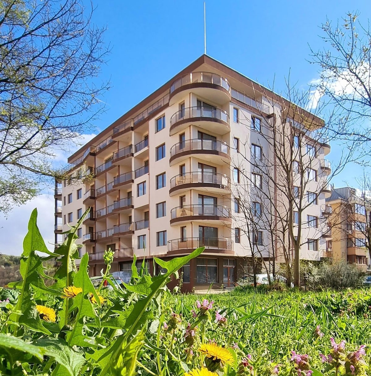 A modern six-story building is surrounded by greenery, featuring multiple balconies that extend from its facade. Natural light illuminates the exterior, highlighting the warm tones of the walls. Flowering plants and grass are visible in the foreground, adding a touch of nature.