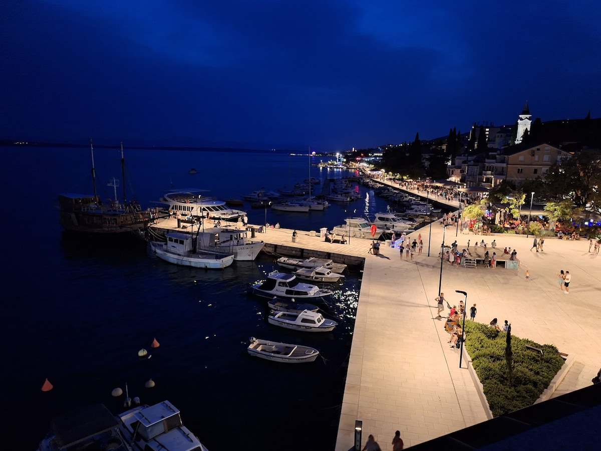 The night scene captures a waterfront area bustling with activity. Boats are docked along the pier, while pedestrians enjoy the promenade lined with lights. The calm water reflects the glow of nearby establishments, creating a vibrant atmosphere against the darkening sky.