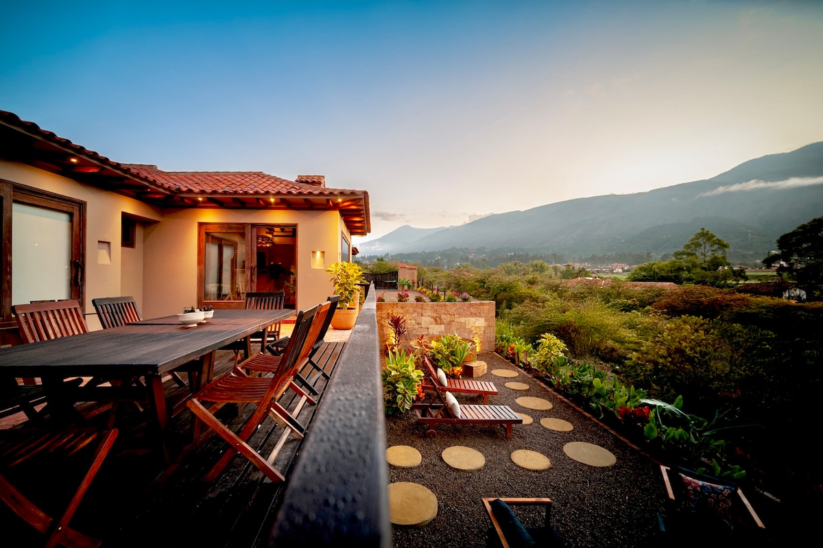 A spacious terrace is shown, featuring a large wooden dining table surrounded by matching chairs. Lush greenery and various plants enhance the outdoor space, while scenic mountain views create a serene backdrop. Soft evening light casts a warm glow over the area.