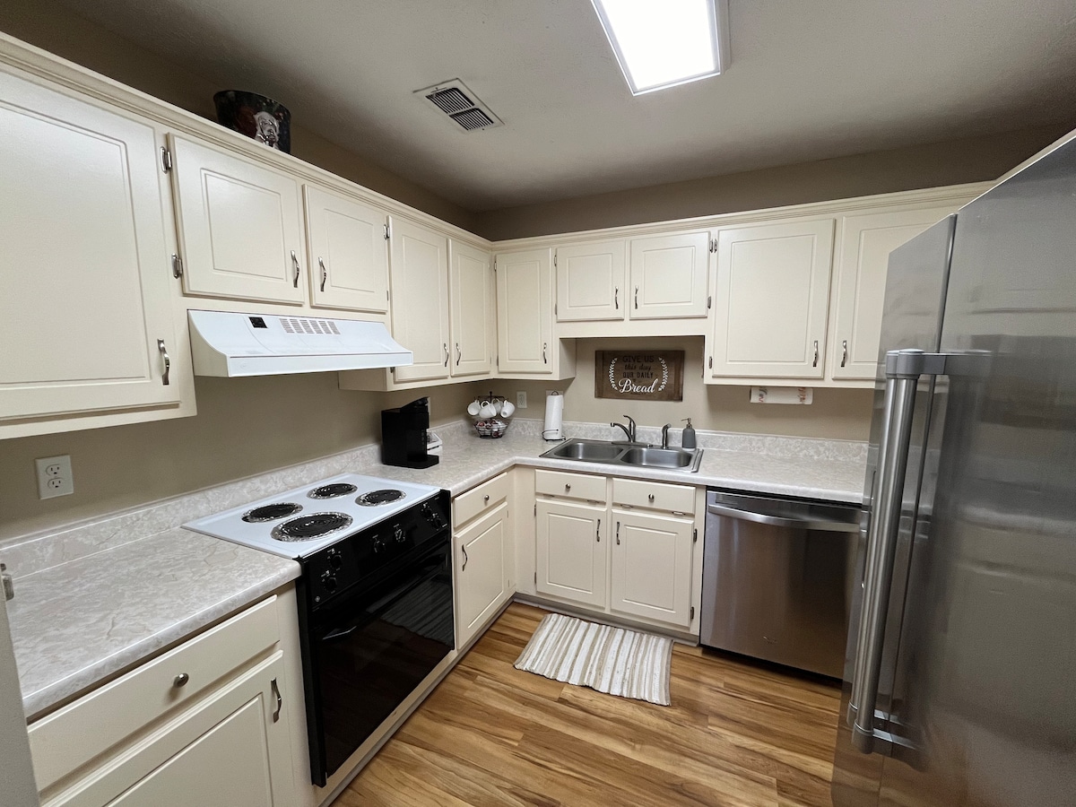 The kitchen features cream-colored cabinetry and light countertops, providing ample space for meal preparation. A black stove and built-in microwave are included, with a stainless steel refrigerator seen nearby. Natural light fills the room from overhead lighting, creating a functional cooking area.