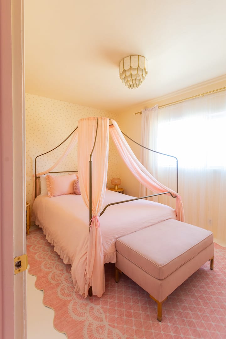 Light-filled butter yellow bedroom with pink chiffon canopy bed, sheer drapes, scalloped rug, and soft neutral tones. Cozy, feminine desert stay near Joshua Tree.