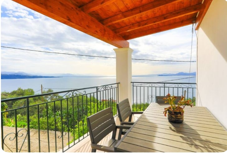 An outdoor terrace features a wooden ceiling with exposed beams. A dining table is set with gray chairs, overlooking scenic views of the coastline and azure waters under a partly cloudy sky.