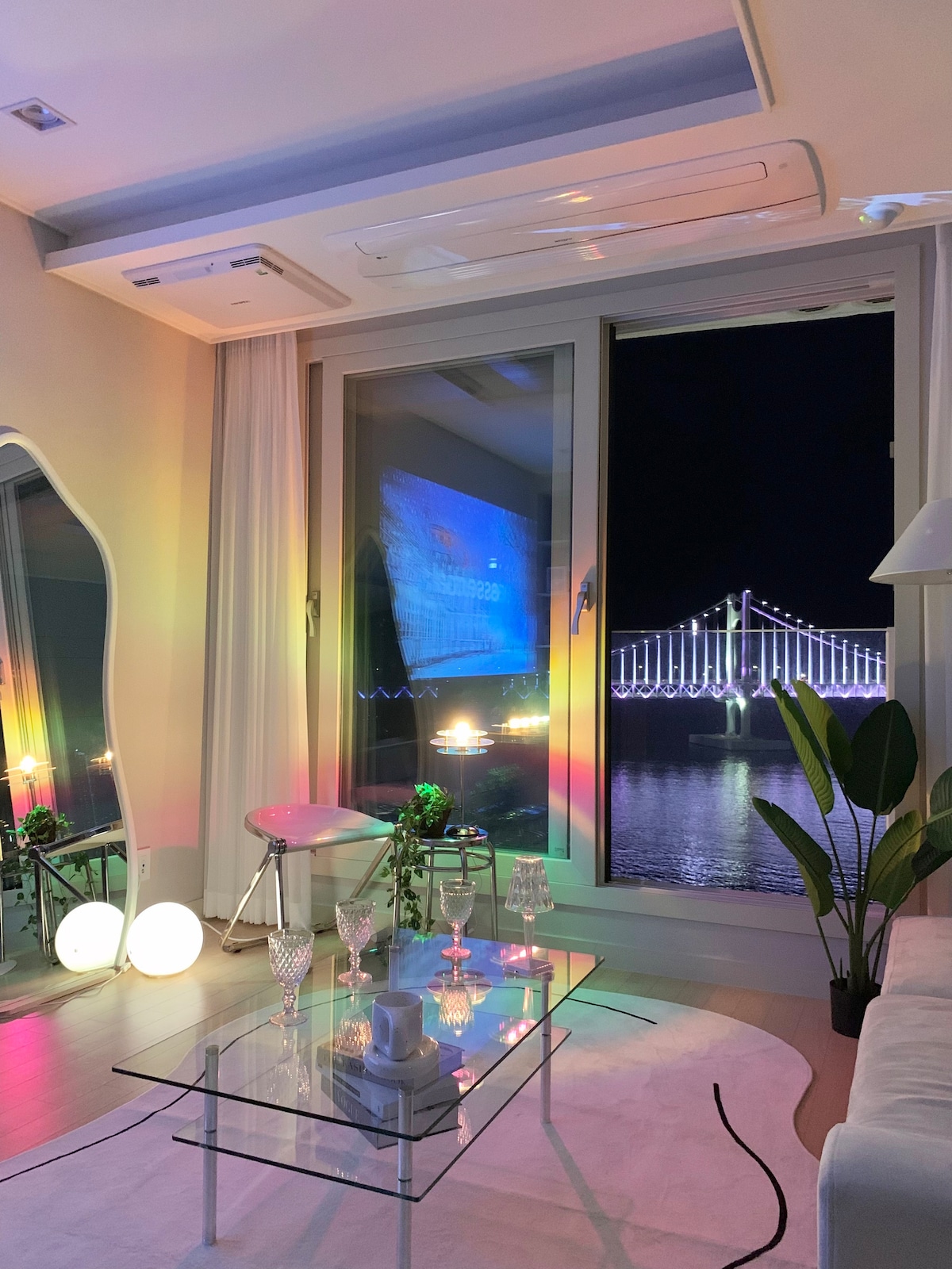 A spacious living area is shown, featuring large windows that provide a view of the Gwangan Bridge illuminated at night. A sleek glass coffee table is set with glassware, while soft lighting creates a soothing ambiance. Indoor plants add a touch of greenery to the space.