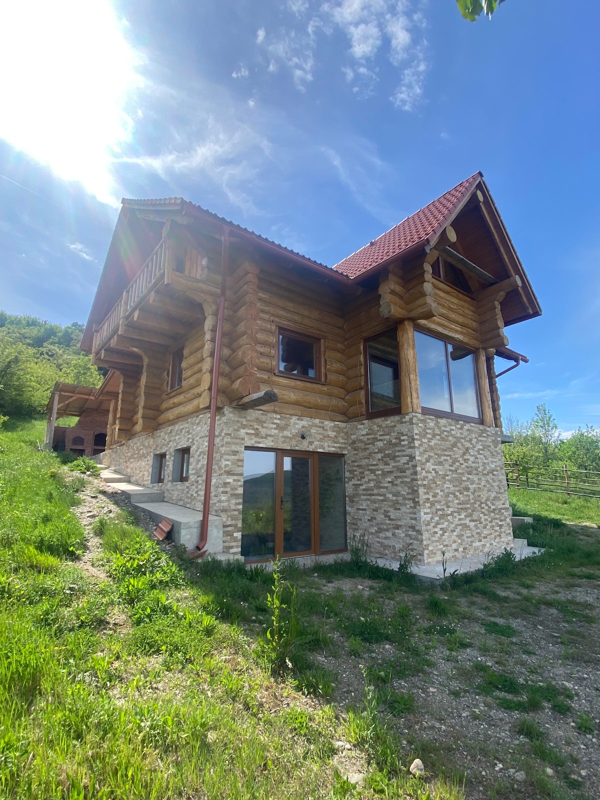 A charming cottage constructed from Szekler wood logs is depicted against a clear blue sky. The building features a sloped red roof and multiple large windows that bring in natural light. Surrounding greenery enhances the natural setting.