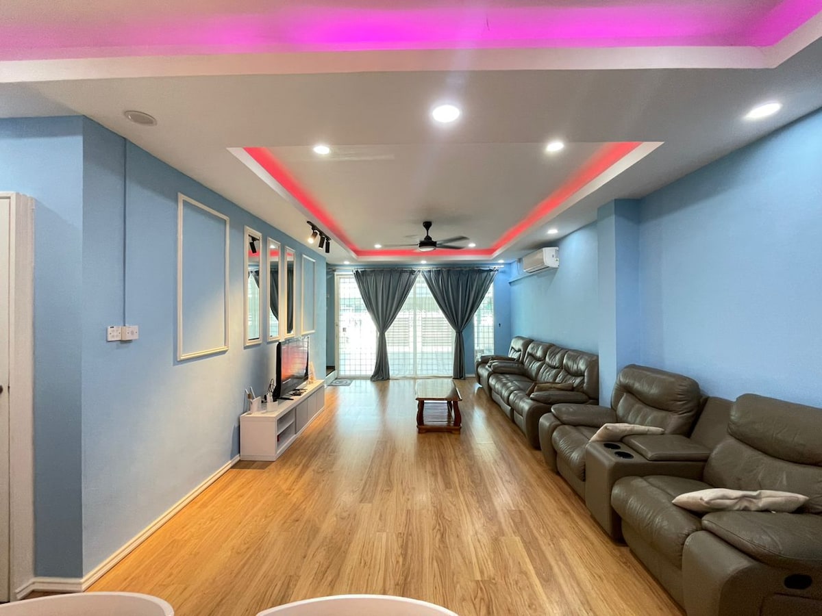 The living room features a spacious layout with light blue walls and warm wooden flooring. Comfortable leather sofas are arranged for relaxation, while a flat screen TV is mounted on the opposite wall. Floor-to-ceiling curtains frame the large windows, allowing natural light to fill the space.