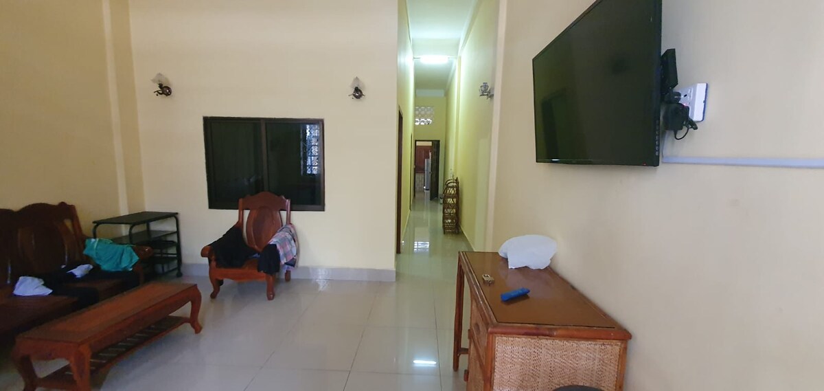 A shared living room is shown, featuring a wooden coffee table and seating arrangements. A wall-mounted television is visible opposite the seating area. The room is well-lit, with a narrow hallway leading to additional spaces, and a large window allowing natural light to enter.