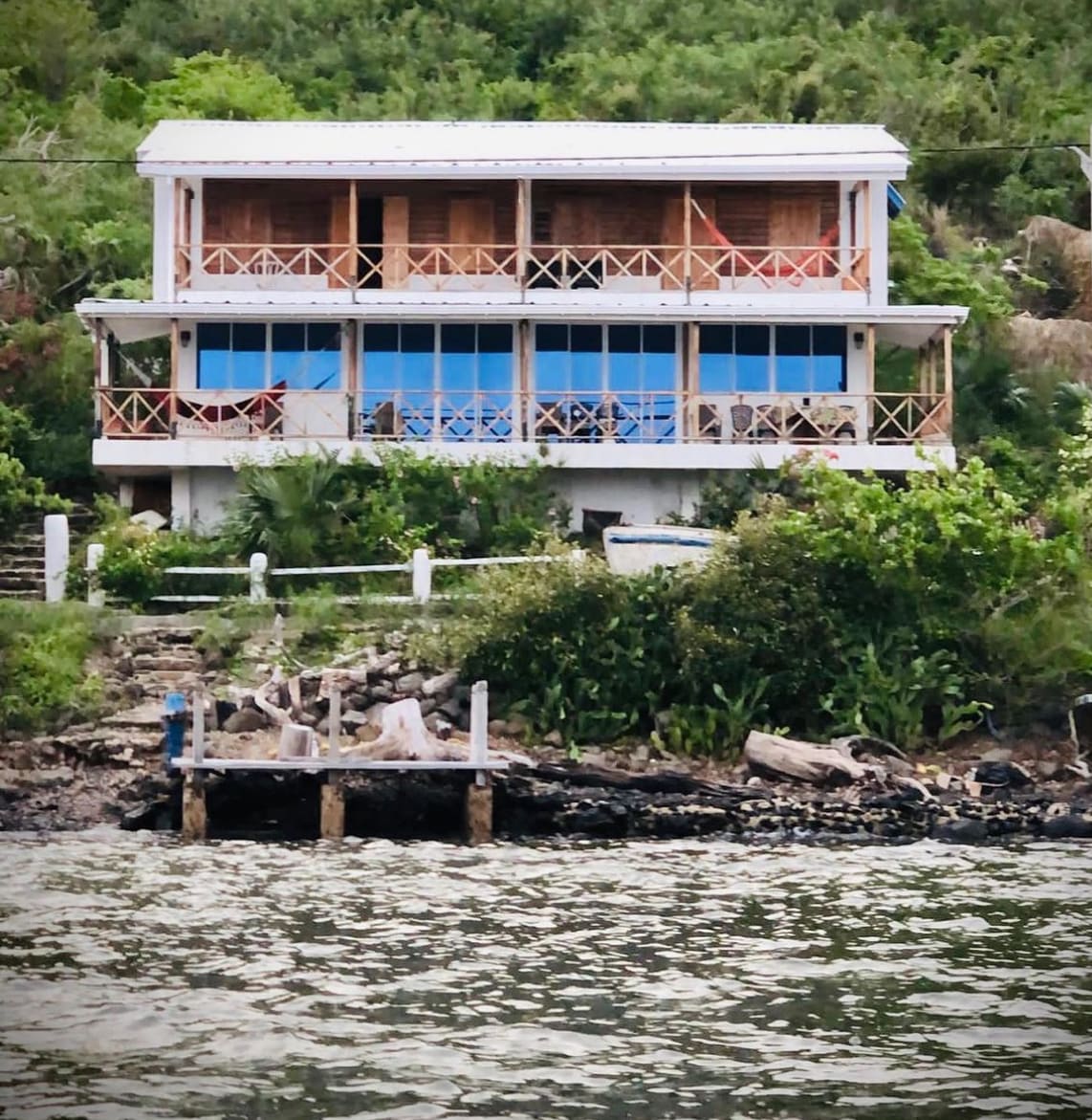 A two-story coastal house is shown, featuring expansive blue windows and a spacious balcony with wooden railings. Surrounding greenery complements the property, while a dock extends into the calm water, providing easy access to the shoreline.