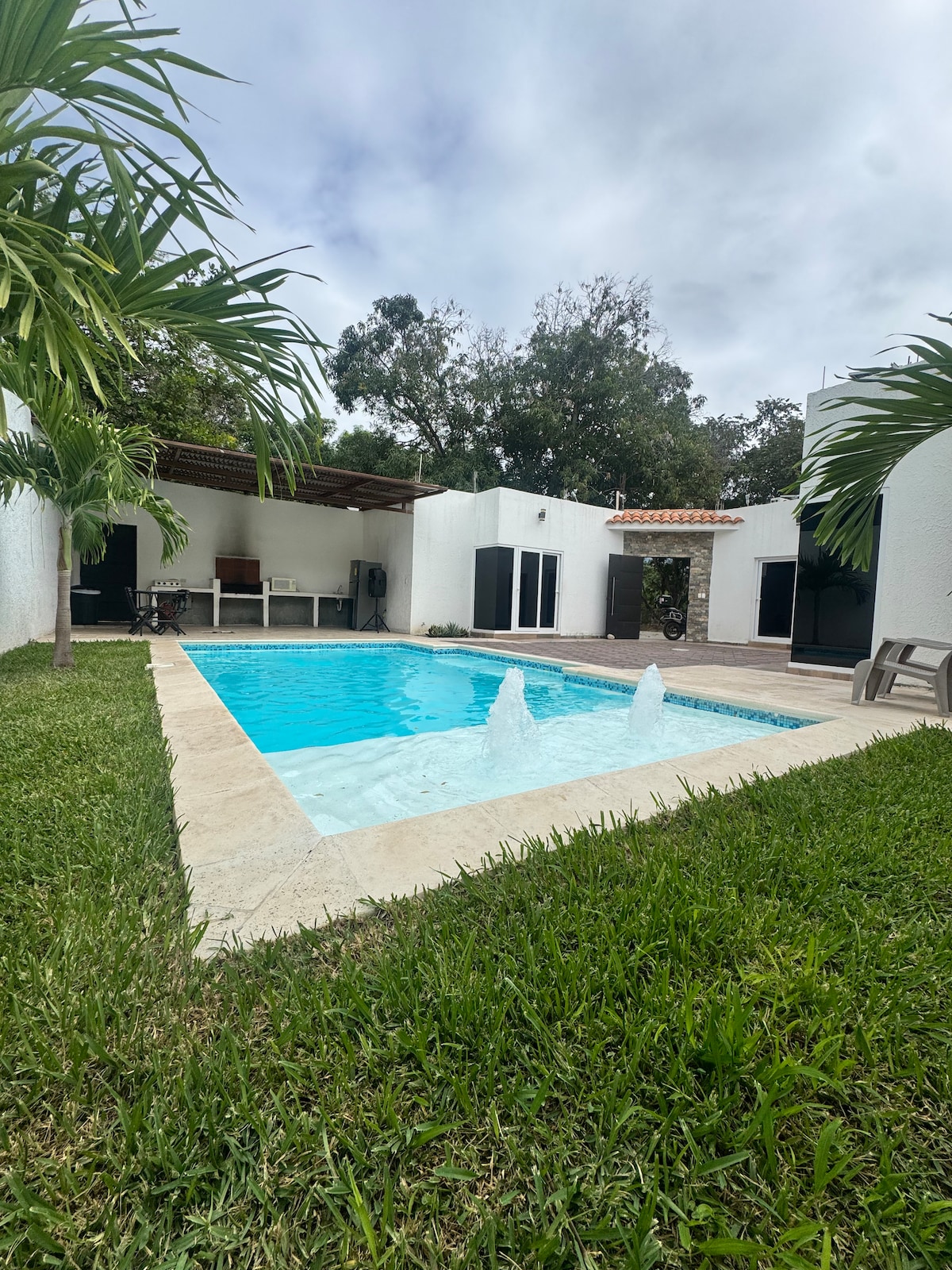 A tranquil pool area surrounded by lush greenery is showcased. The inviting water features three jets, while lounge chairs are positioned for relaxation. The neighboring structures are designed with large windows, integrating nature with the exterior spaces. A shaded patio area offers additional seating.