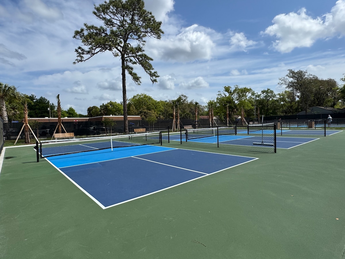 The image showcases multiple tennis courts with blue and green surfaces. Lush trees are visible in the background, providing shade and natural beauty. Clear skies and scattered clouds create a bright atmosphere. The space appears well-maintained, ideal for recreational activities.