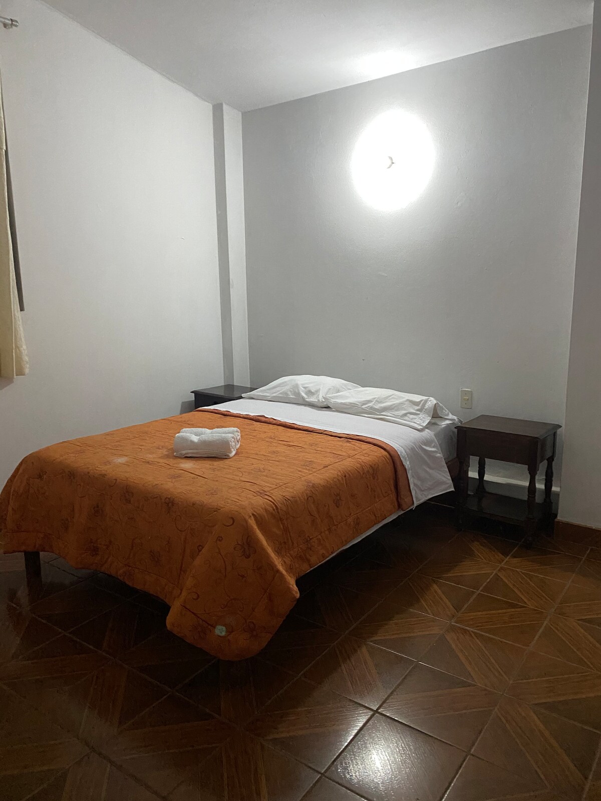 A modest bedroom features a spacious bed covered with an orange blanket. A white towel is neatly placed on the bed. Two nightstands are positioned beside the bed, and soft lighting from a wall fixture creates a calm ambiance in the area.