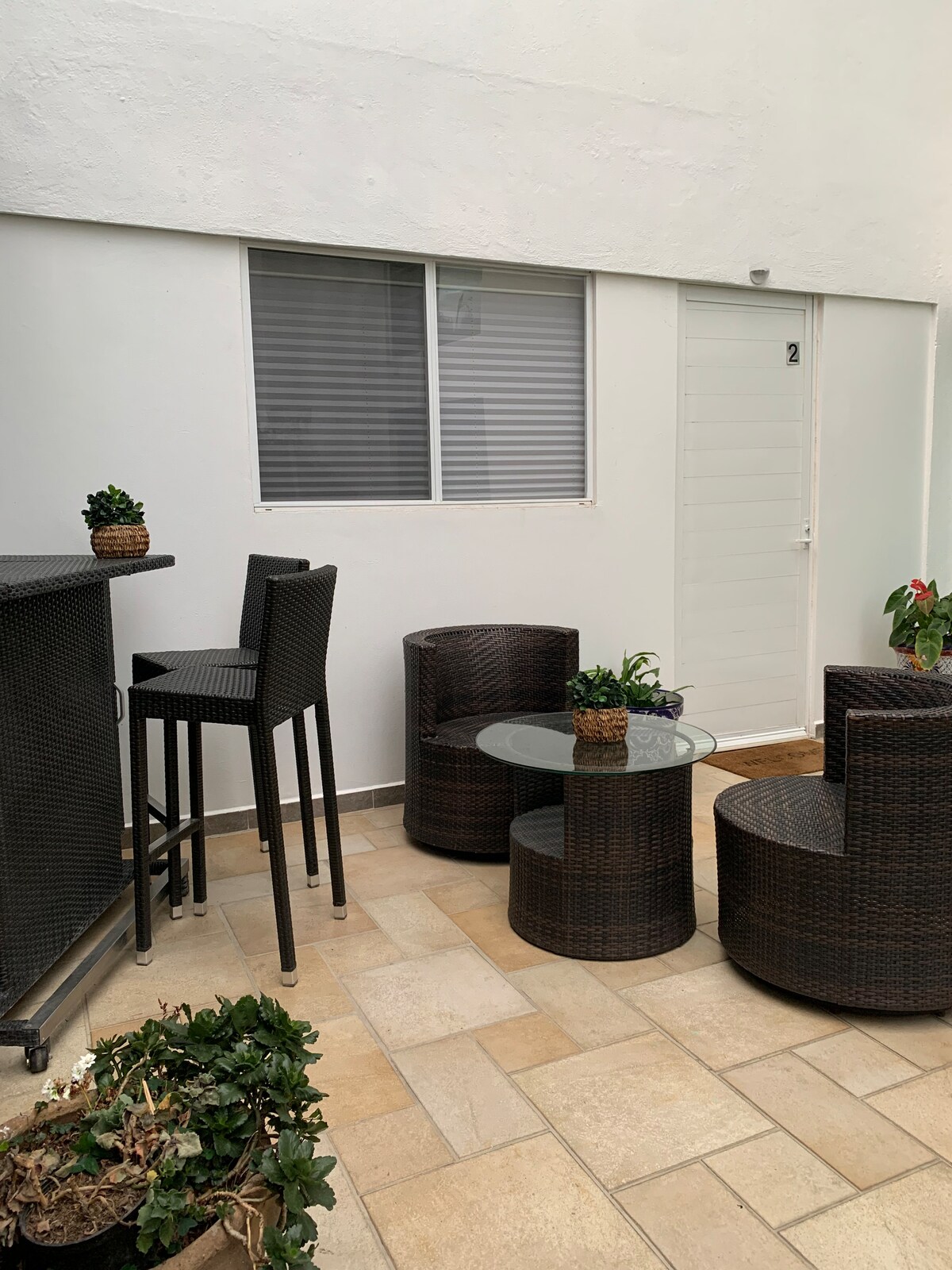 An outdoor sitting area features modern dark wicker furniture, including a glass top table and two circular seating arrangements. A tall bar table with stools is present at one side. Potted plants and a door marked '2' provide a welcoming touch to the space.