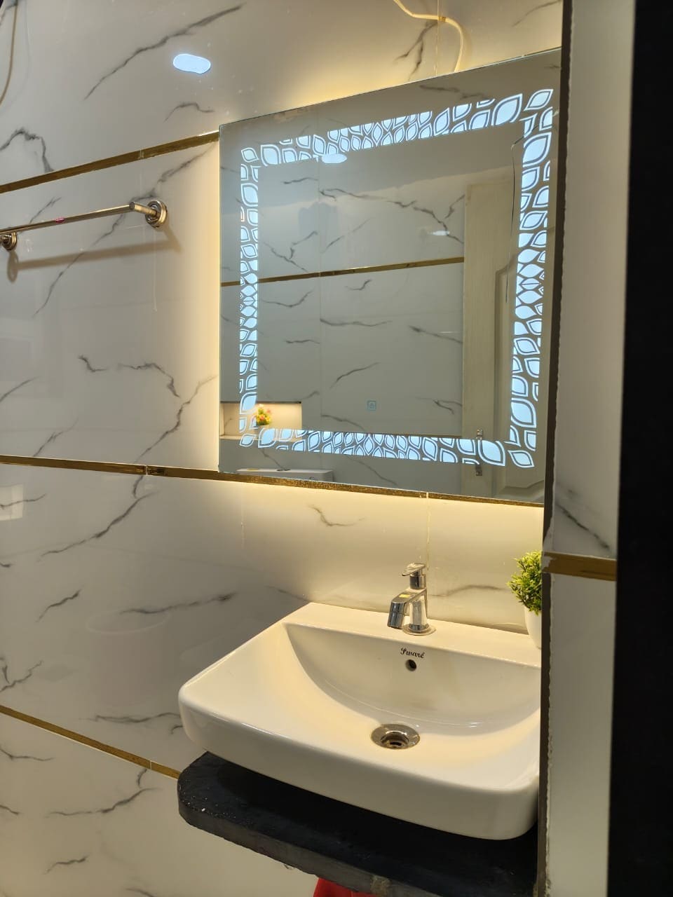 A modern bathroom sink area is presented, featuring a sleek white basin with a single faucet. A well-lit, decorative mirror reflects the stylish geometric design, while subtle marble patterns adorn the walls. A small plant adds a touch of greenery to the space.