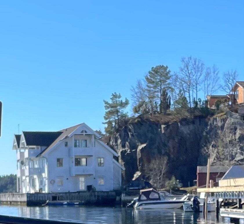 A two-story white building is situated by the water, featuring a porch and multiple windows. A rocky cliff covered with trees rises in the background. Several boats are visible in the water, suggesting an active maritime environment.