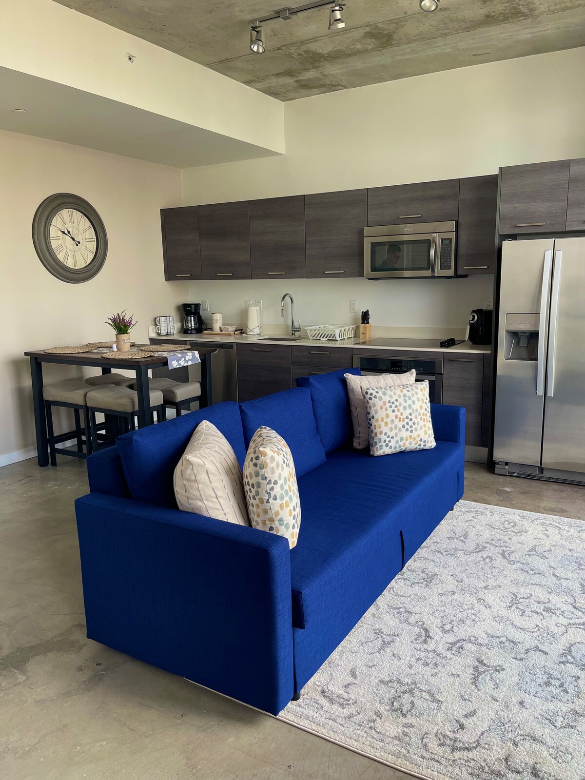 A bright and functional living area features a blue sofa adorned with decorative pillows. A dining table with chairs is visible nearby, along with a well-equipped kitchen showcasing modern cabinetry and appliances, including a microwave and refrigerator. Natural light enhances the space.