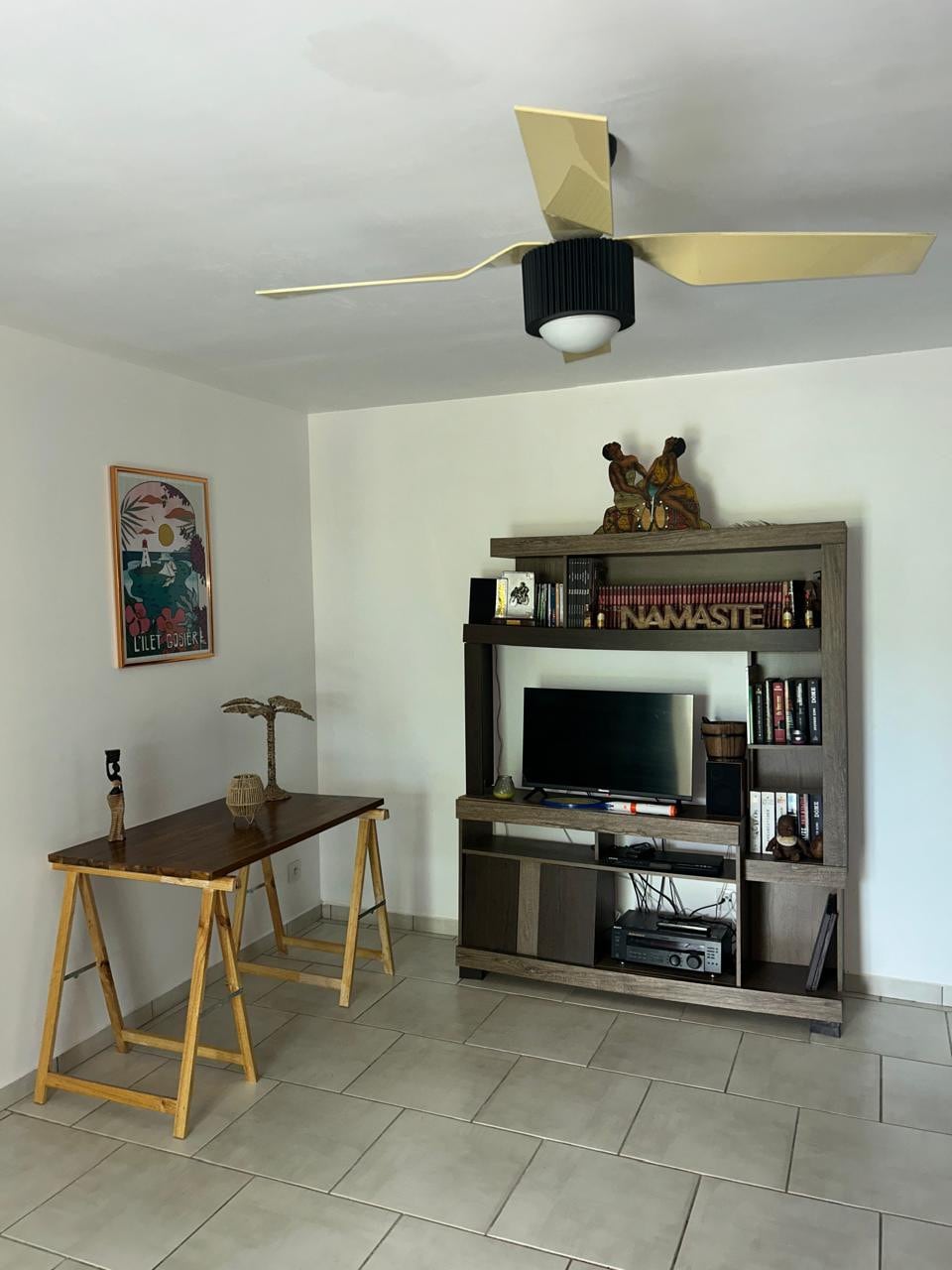 A living area features a wooden desk with a minimalist design, accompanied by a decorative table lamp. A stylish bookshelf is positioned against the wall, holding a television and a collection of books. Artwork is displayed on the adjacent wall, enhancing the space.