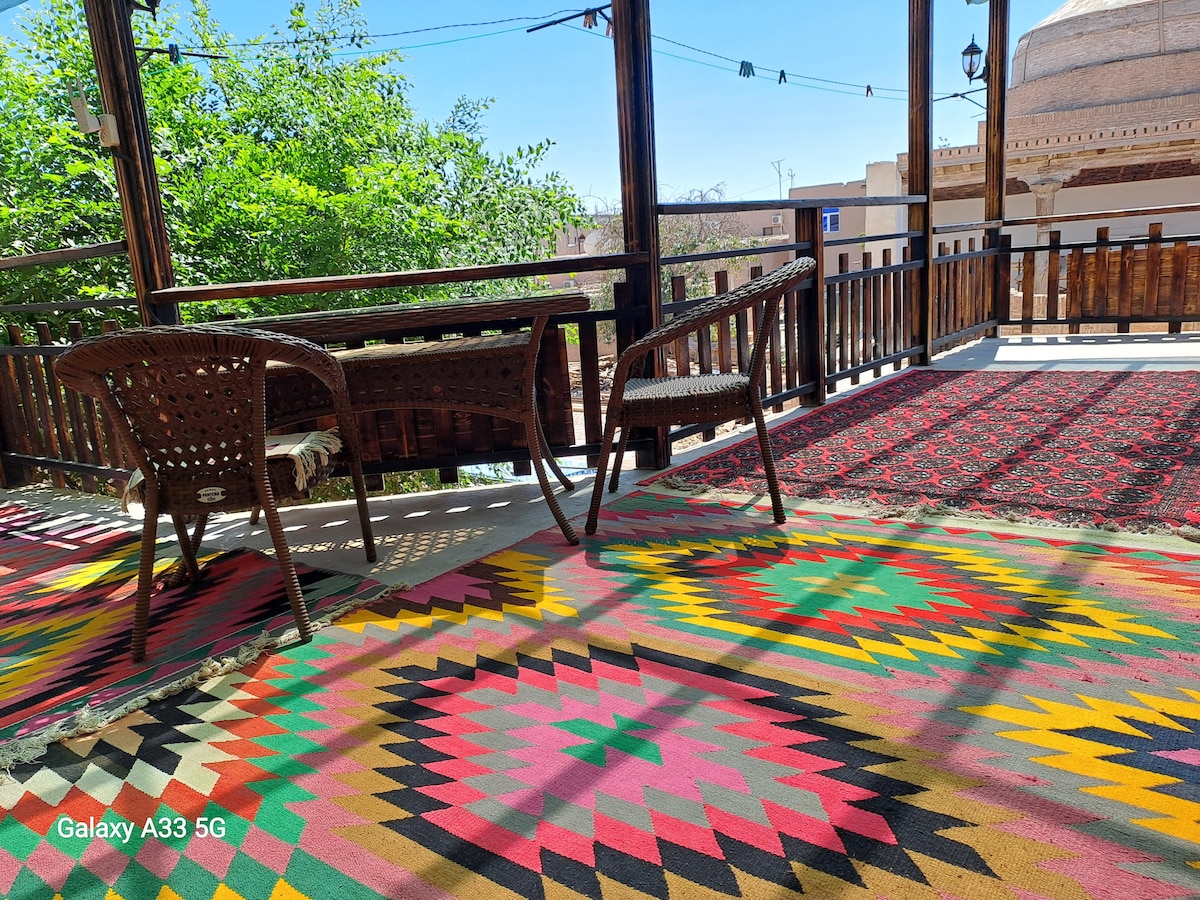 A patio area is featured with two woven chairs positioned around a wooden table. A vibrant, patterned rug in bright colors covers the floor, enhancing the inviting atmosphere. Green foliage surrounds the space, providing a touch of nature.
