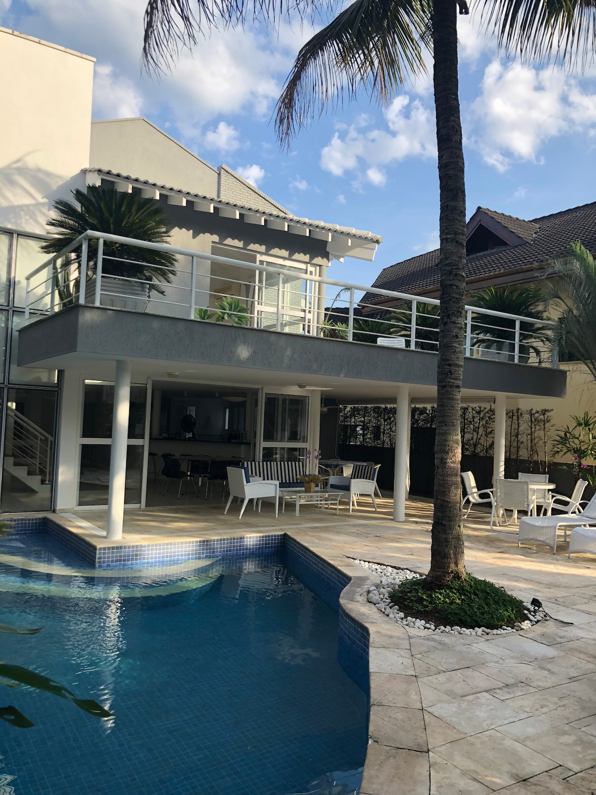 A spacious pool area is framed by a well-maintained garden, featuring a clear blue pool. The two-story house showcases a modern design with a balcony and large glass windows. Several white lounge chairs are arranged around the pool, enhancing the outdoor relaxing space.