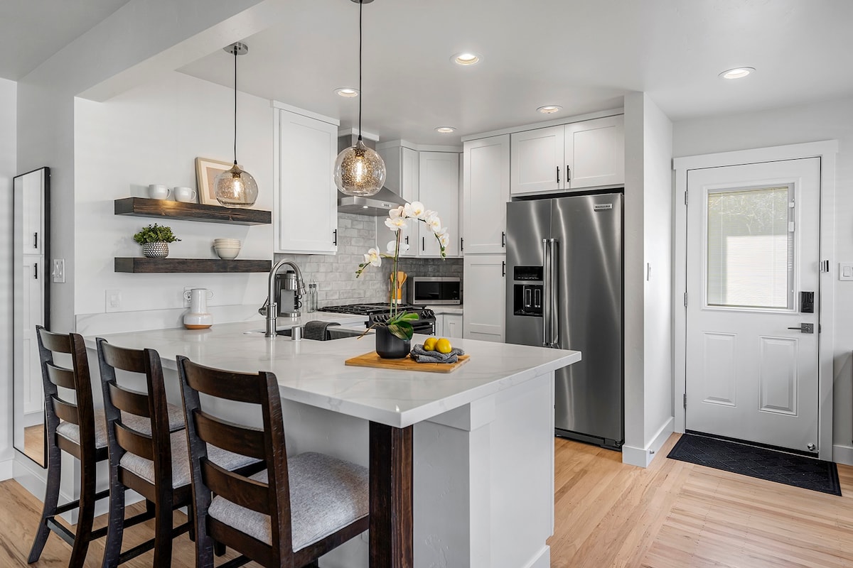 A modern kitchen is presented, featuring stainless steel appliances and a large island with seating for four. Pendant lights hang above the island, and decorative shelving is mounted on the wall. Natural light enters through a nearby door, enhancing the bright atmosphere.