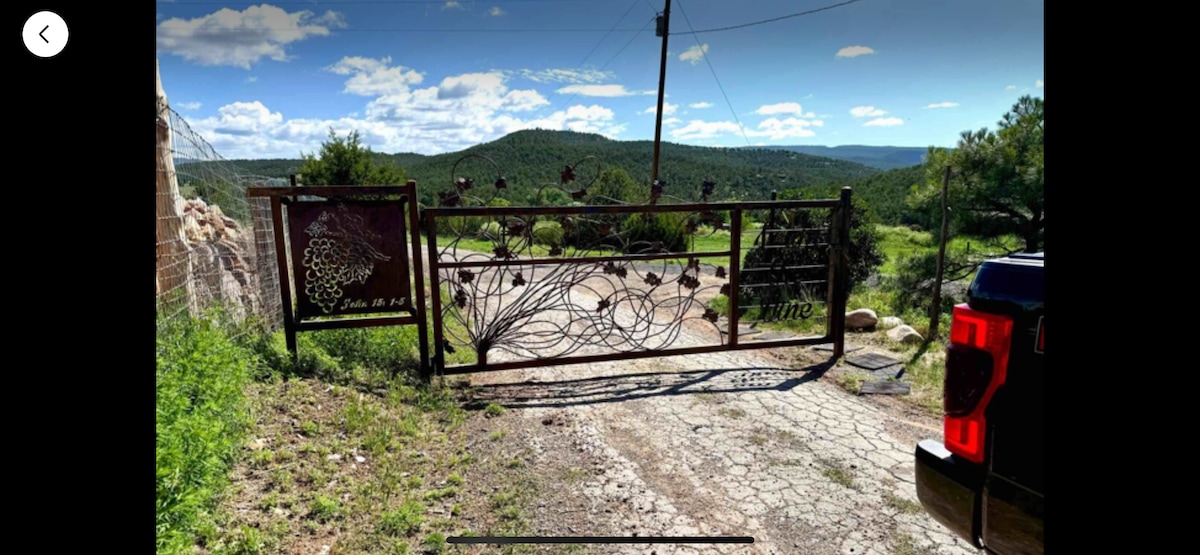 A rustic gate with intricate wrought iron design stands at the entrance, surrounded by lush greenery and a gravel path. Rolling hills are visible in the background under a clear blue sky, creating a serene atmosphere.