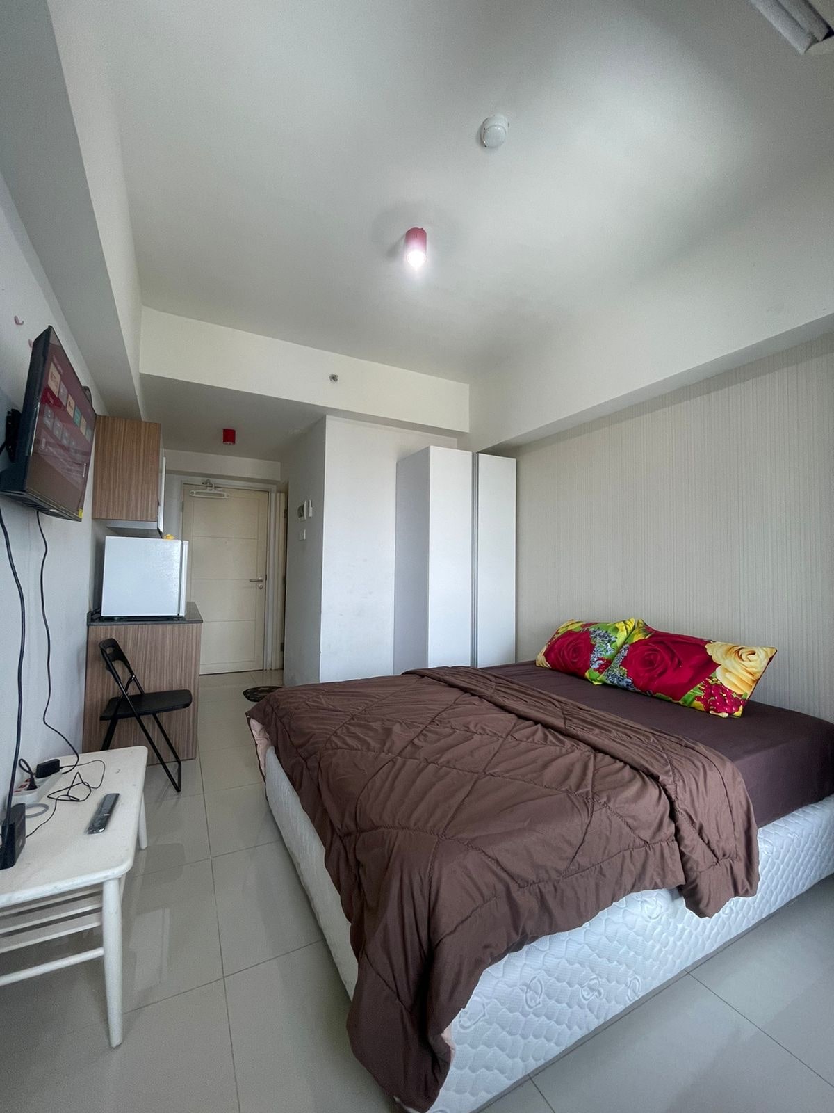 A studio apartment is visible, featuring a king-size bed with a brown blanket and decorative throw pillows adorned with floral patterns. A small TV and refrigerator are positioned against one wall, while a chair and desk are arranged nearby. The space is brightly lit, highlighting the clean, tiled floor.