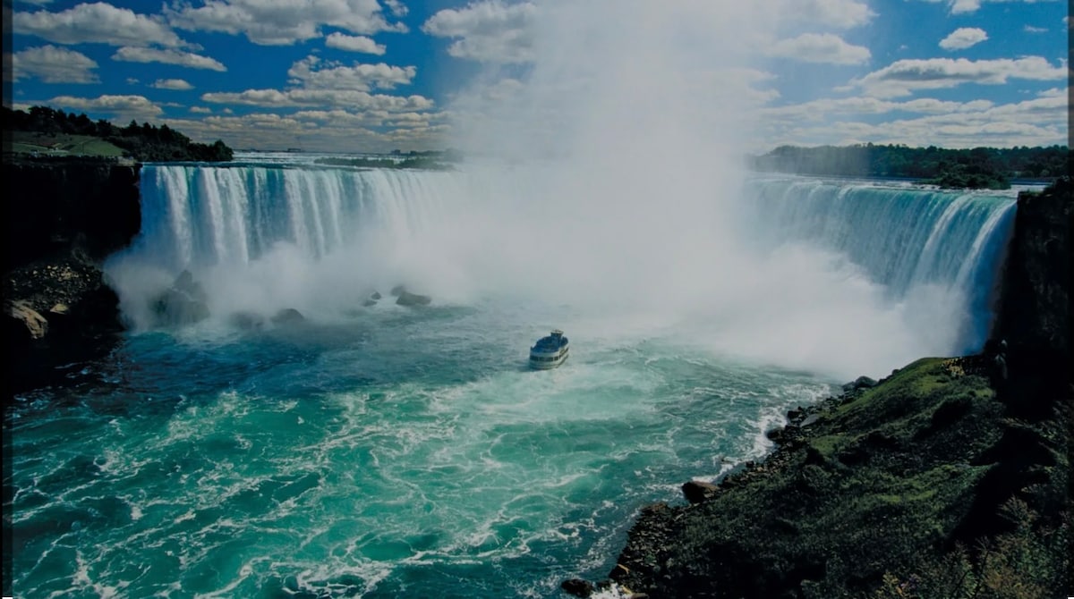 A panoramic view of Niagara Falls displays cascading water pouring down rugged cliffs into a vibrant blue river below. A boat is visible on the water, providing a sense of scale to the powerful natural scenery framed by a partly cloudy sky.