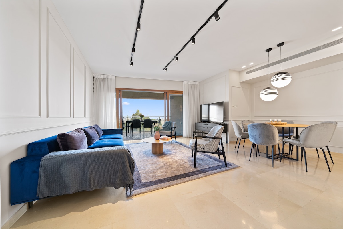 A modern living area showcases a blue sectional sofa and a light wood coffee table. Large glass doors open to reveal a view, with a dining area featuring a round table and black chairs situated nearby. The space is illuminated by sleek pendant lights.