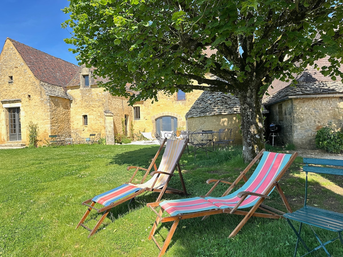 A spacious outdoor area is presented, featuring wooden lounge chairs with striped fabric positioned beneath a large tree. The rustic stone house is visible in the background, accompanied by additional seating and decorative elements scattered throughout the grassy lawn.