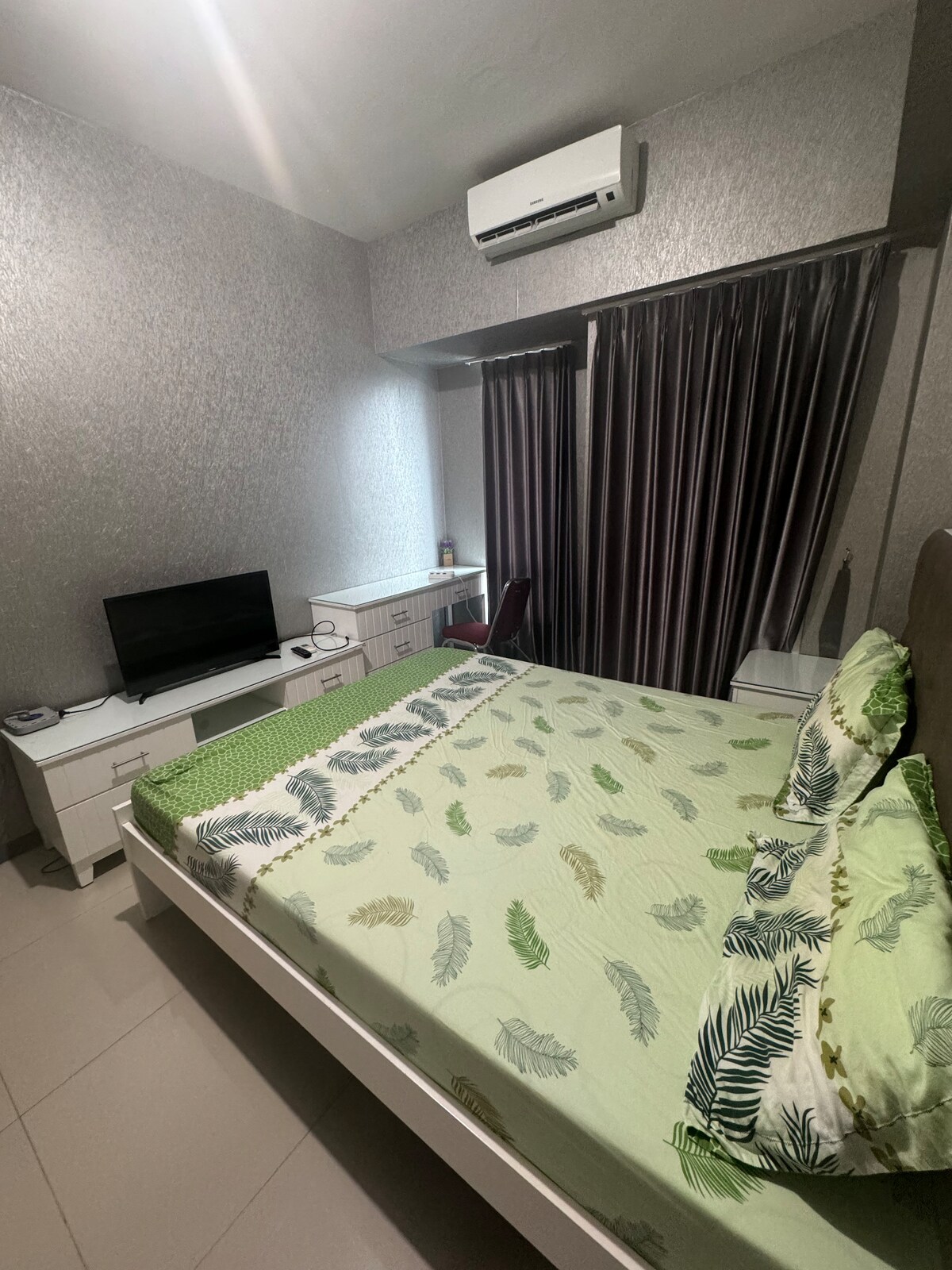 A well-appointed bedroom features a spacious bed with a patterned green and white duvet. A television is positioned on a white cabinet, accompanied by a desk and chair. Natural light filters through a window, which is framed by dark curtains.