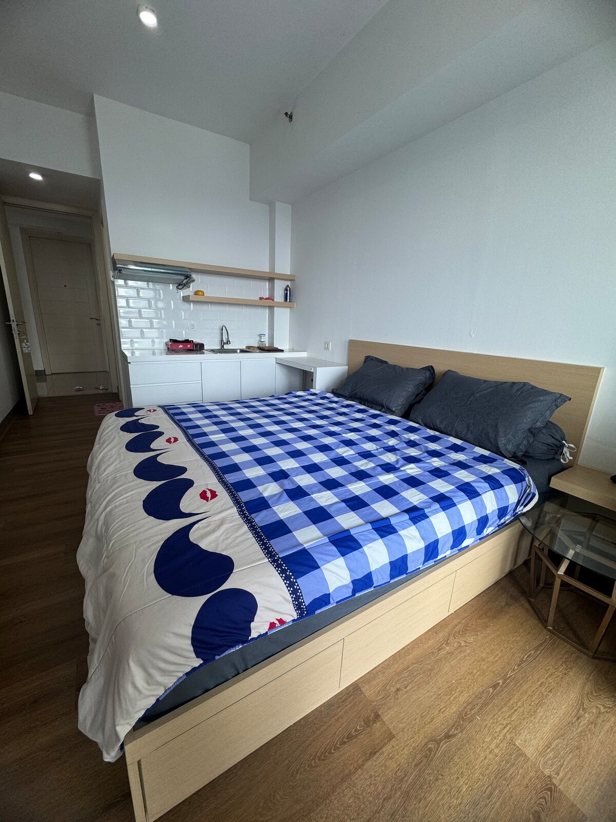 A spacious bedroom features a double size bed dressed in a blue and white checkered blanket, complemented by dark grey pillows. A kitchenette is visible in the background, equipped with essential appliances and cabinetry. Light wood flooring adds warmth to the space.