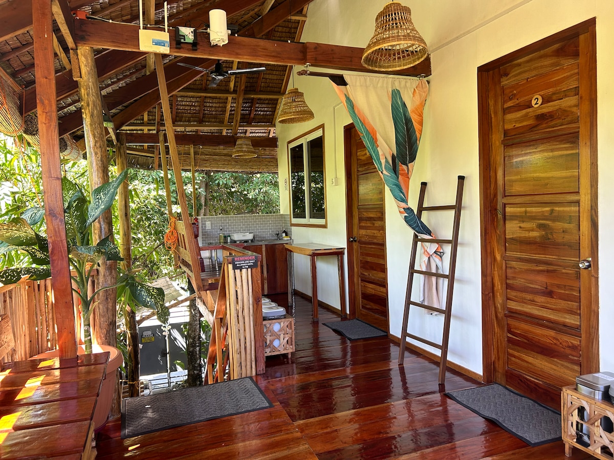 A spacious outdoor area features wooden flooring and a natural wood ceiling. A kitchen space is visible in the background, equipped with cabinetry. Two doors lead to individual rooms, while a decorative ladder and tropical-themed fabric add a touch of color. Lush greenery surrounds the space.