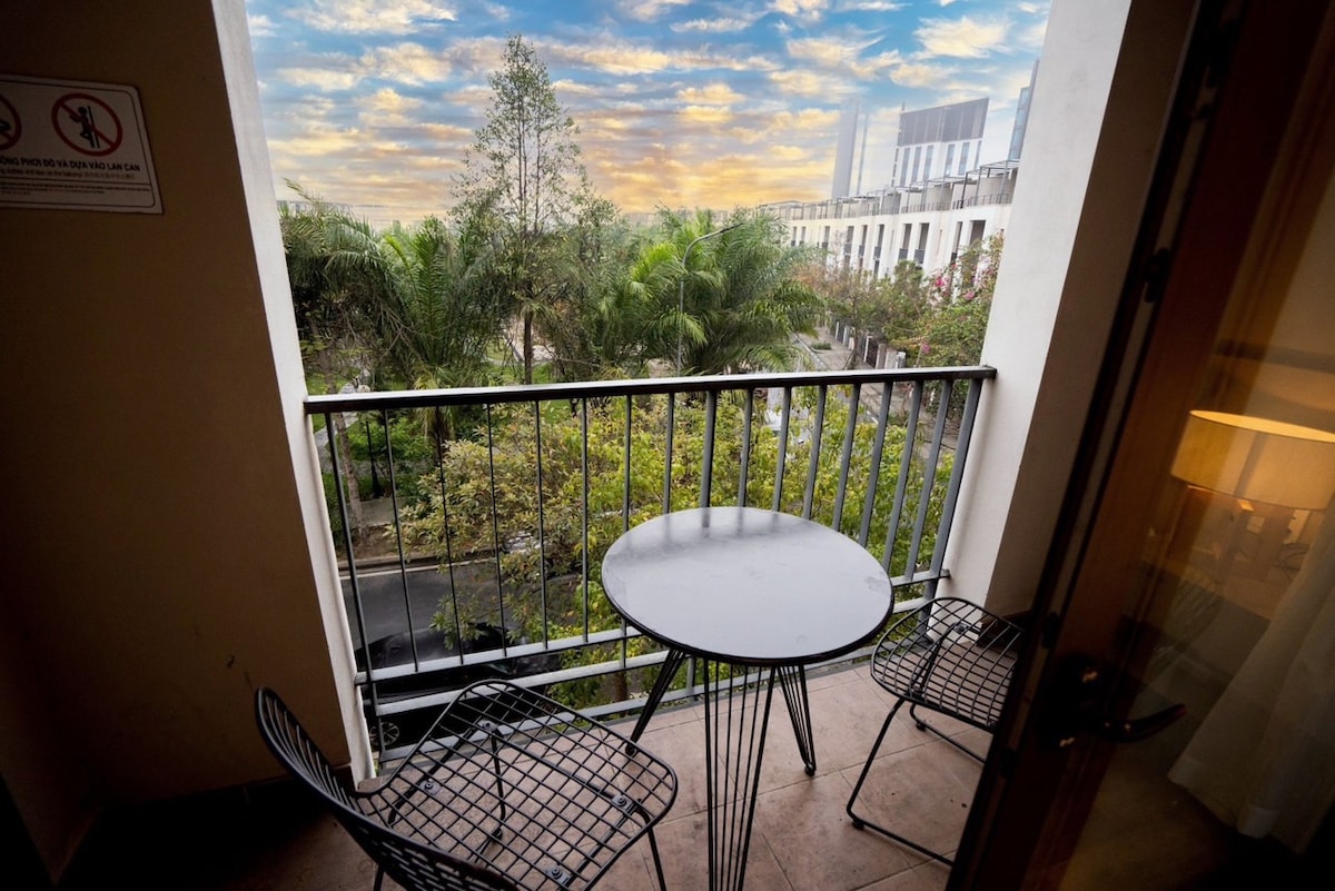 A balcony offers a circular table accompanied by two sleek black chairs. Lush green trees are visible below, along with a glimpse of the street. The backdrop reveals a cloudy sky with warm tones, providing a serene outdoor space.