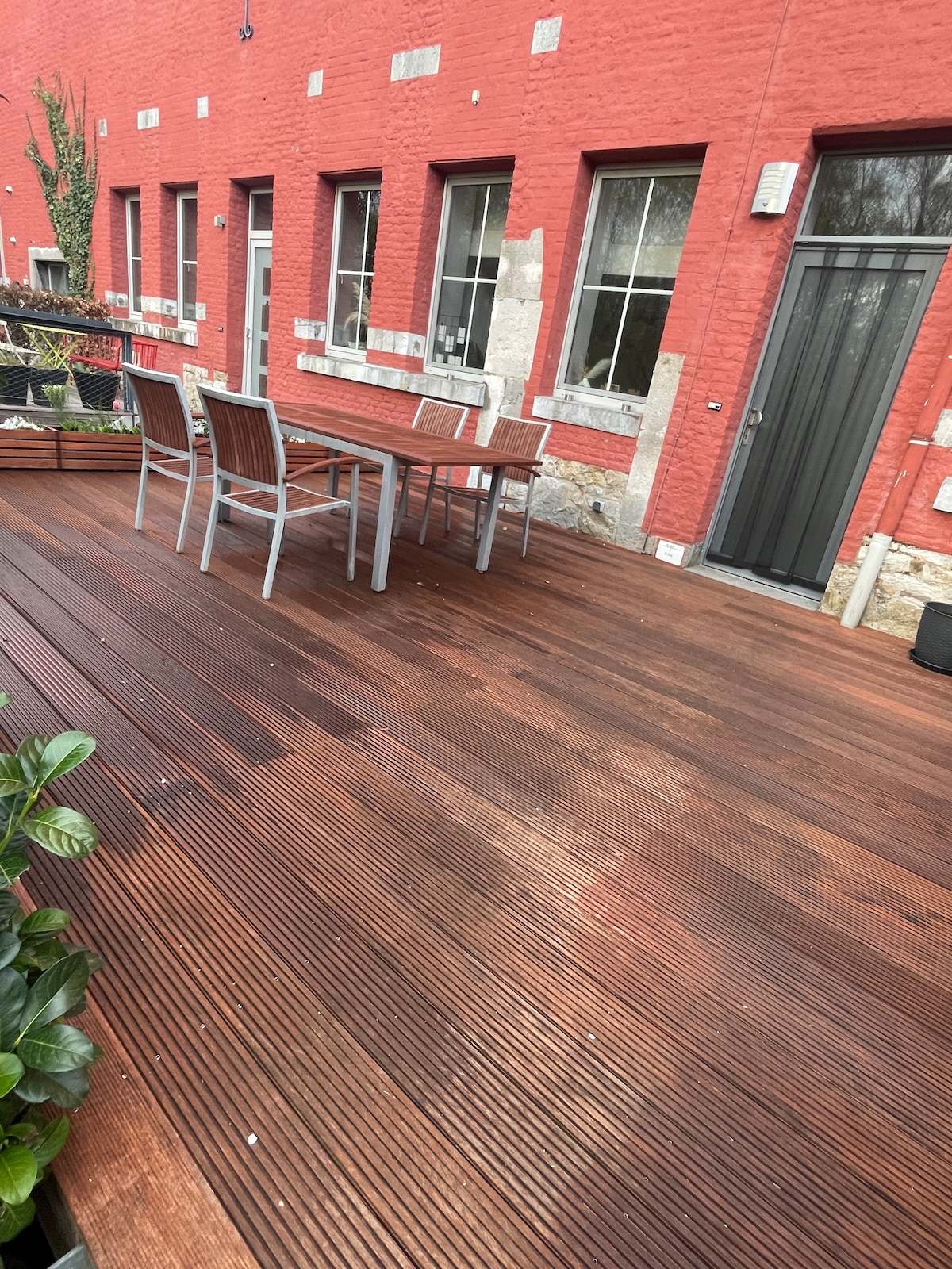 A wooden deck area features a large outdoor table with matching chairs, surrounded by greenery. Large windows of a red brick building are visible, enhancing the space's natural light and inviting ambiance.