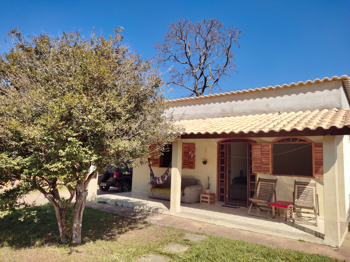A charming house is presented with a tiled roof and wooden shutters. A shady tree adorns the front yard, accompanied by patio seating for relaxation. The entrance features a cozy hammock, inviting guests to unwind in the serene outdoor environment.