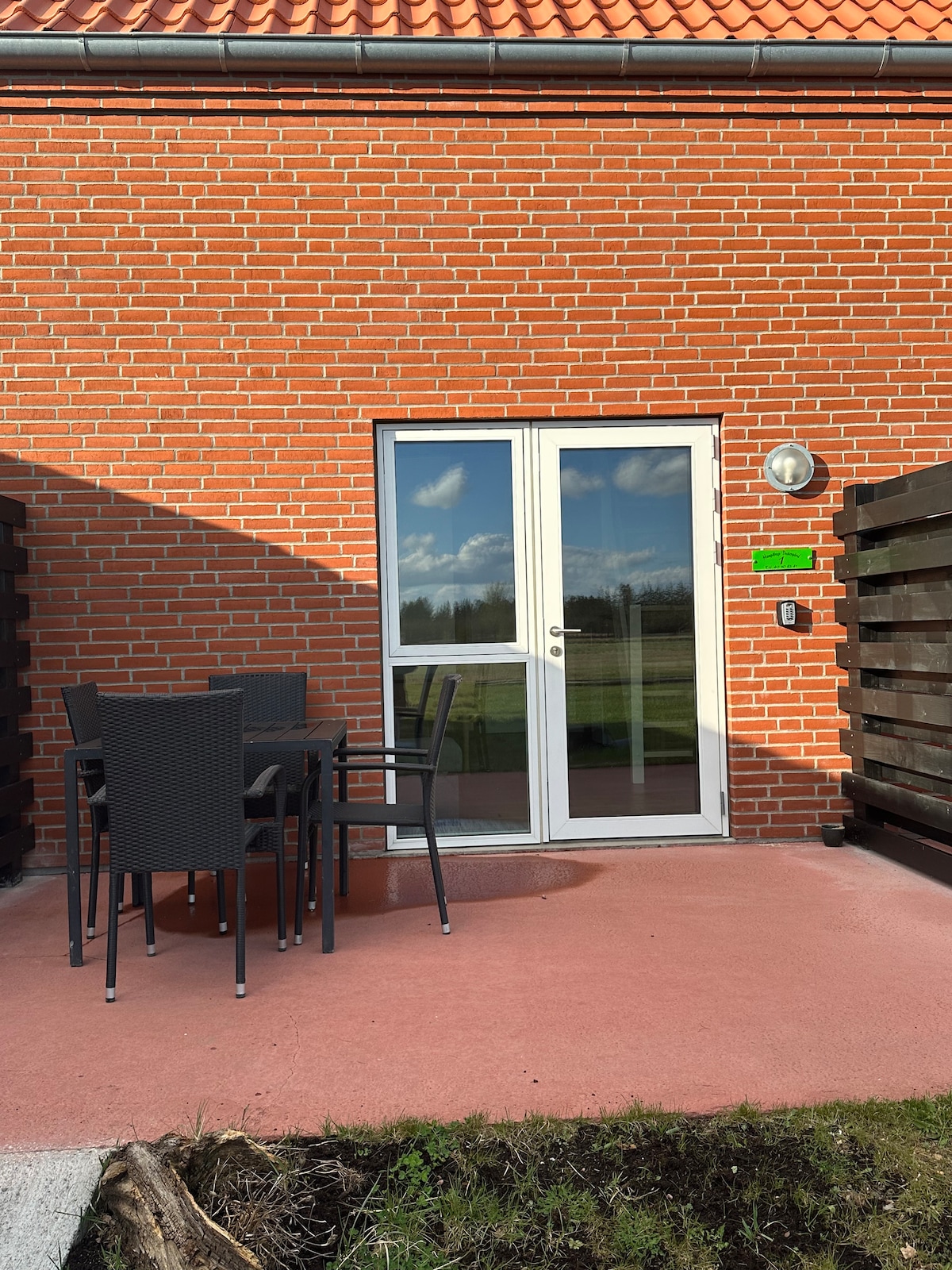 An outdoor patio area is displayed, featuring a set of sleek black chairs and a table. A pair of glass doors leads inside, reflecting the greenery beyond. The brick wall presents a warm contrast, enhancing the sense of openness to nature.