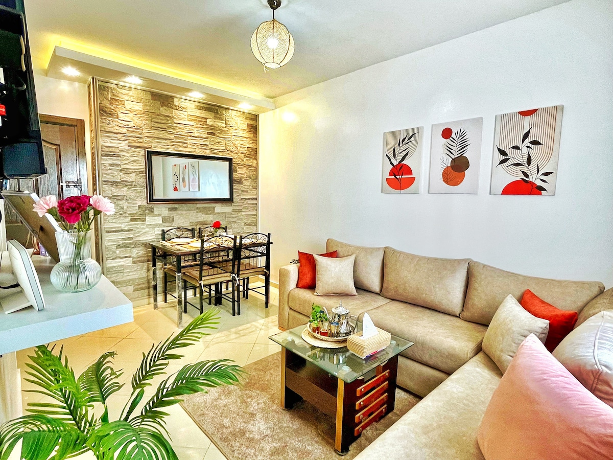A modern living area features a comfortable corner sofa adorned with decorative cushions in warm tones. A small coffee table is set with decorative items. Behind, a dining area is visually separated by a stone accent wall, with a table and chairs for four, complemented by artwork on the walls.