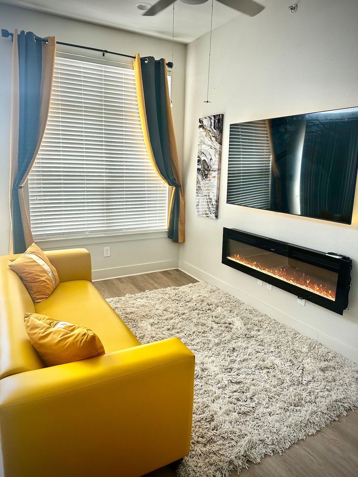 A bright living area is highlighted by a plush yellow sofa positioned on a soft area rug. A mounted flat-screen television and an electric fireplace create a cozy focal point. Soft natural light filters through large windows adorned with blackout curtains.