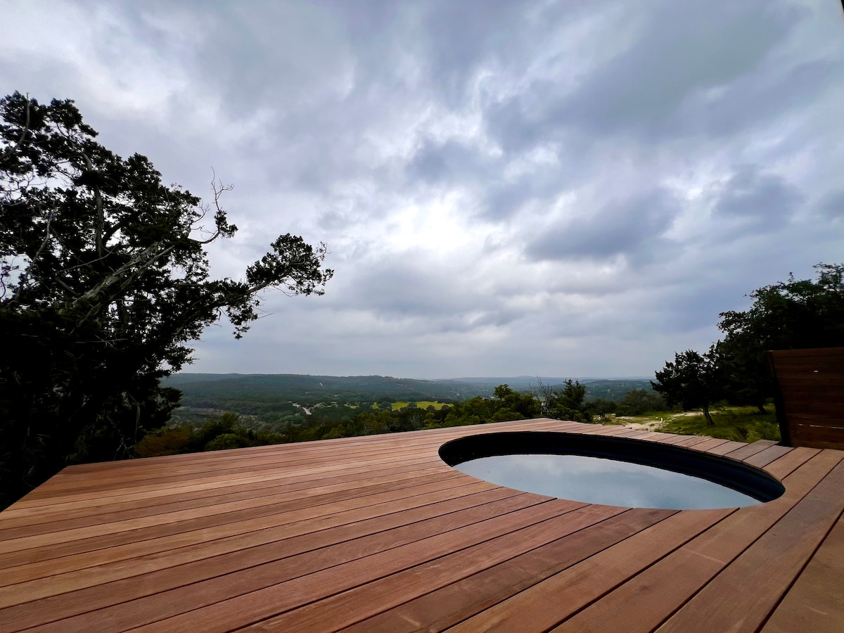 A private hot tub is situated on a spacious wooden deck, offering expansive views of the Texas Hill Country. The grey sky complements the serene atmosphere, while natural vegetation frames the scene, providing a tranquil, secluded setting for relaxation.