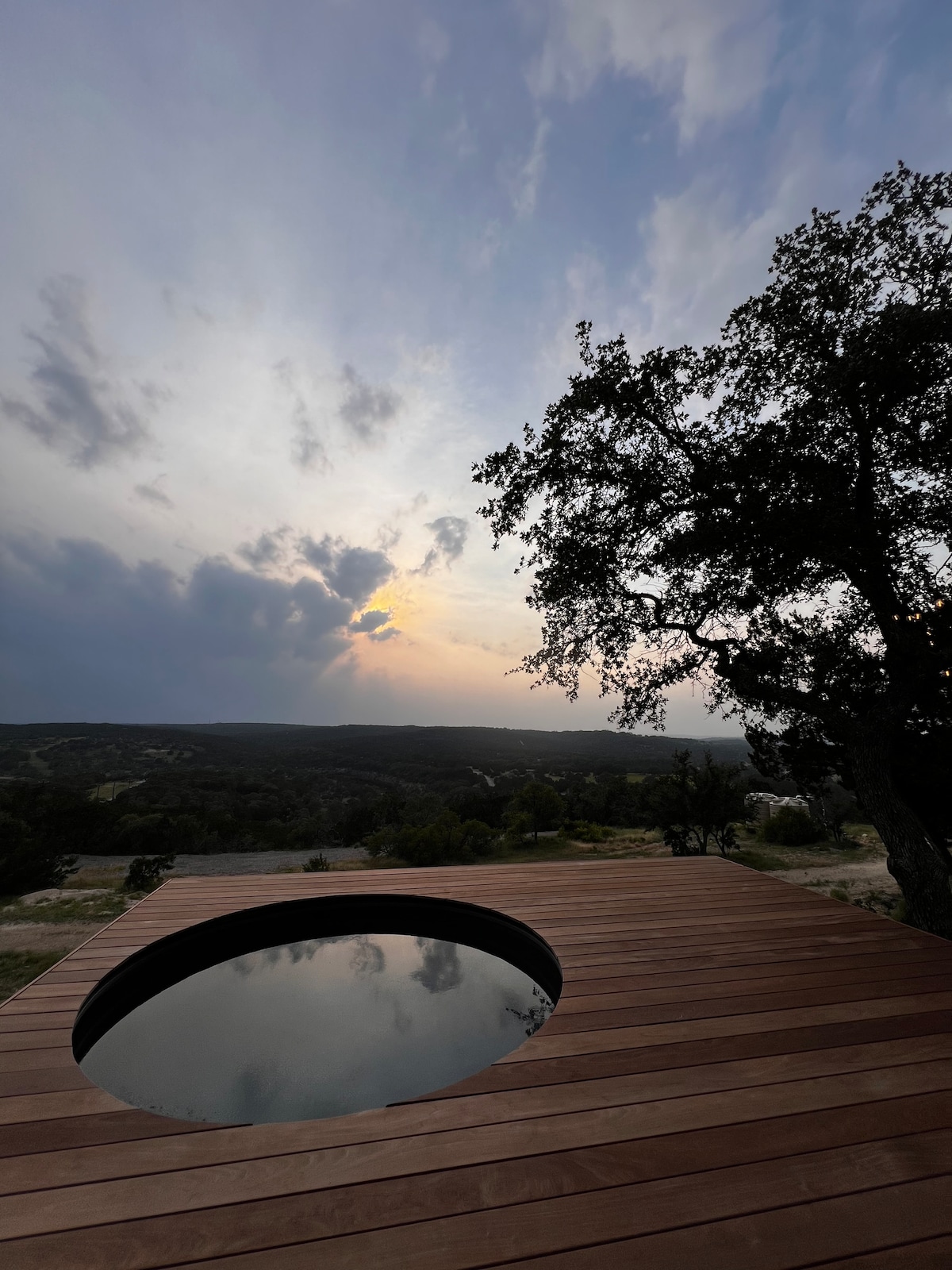 An expansive wooden deck surrounds a private hot tub, reflecting the cloudy sky and upcoming sunset. Lush trees frame the view, offering a serene backdrop of the Texas Hill Country and Flat Creek below.