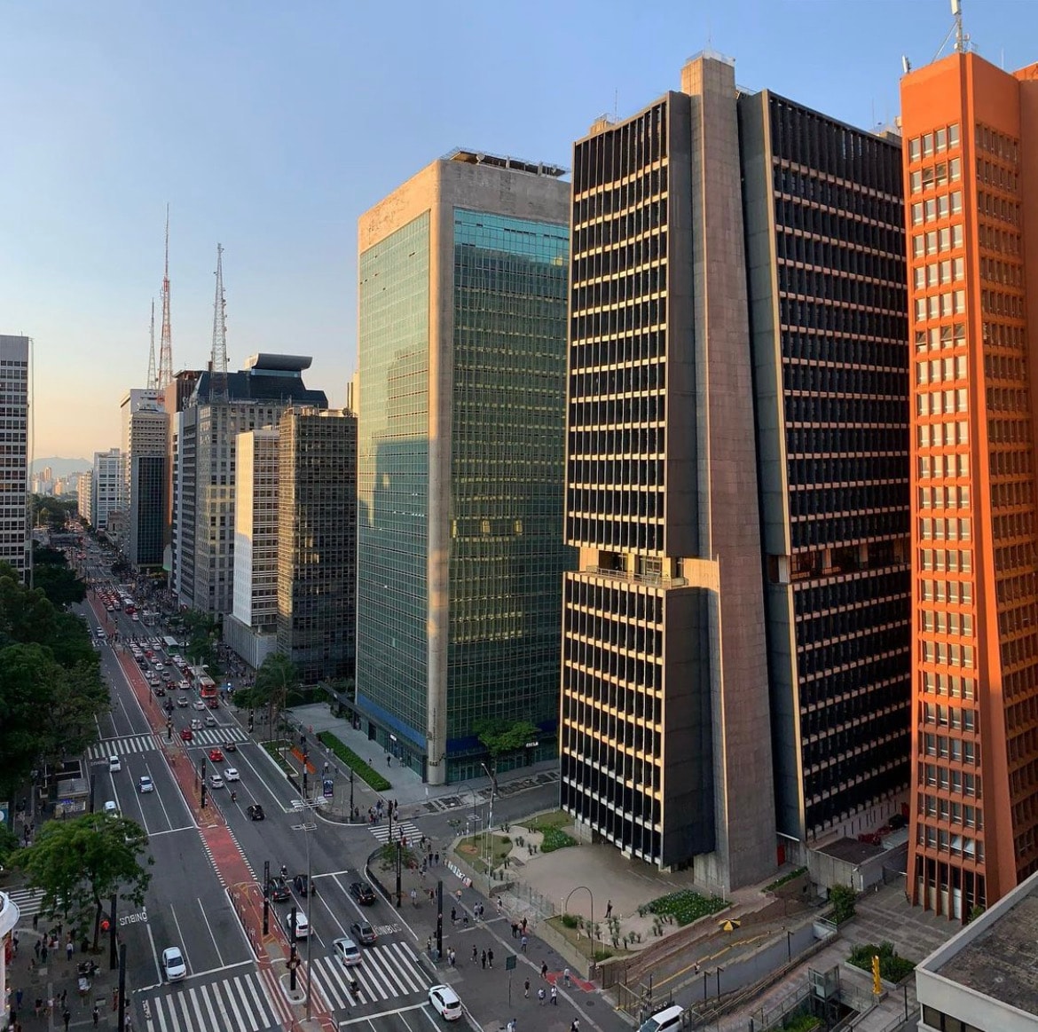 A view from above shows Avenida Paulista, lined with a mix of modern and historic buildings. The busy street is filled with vehicles and pedestrians. Green trees are seen along the sidewalks, with the city skyline prominent in the background under a clear sky.