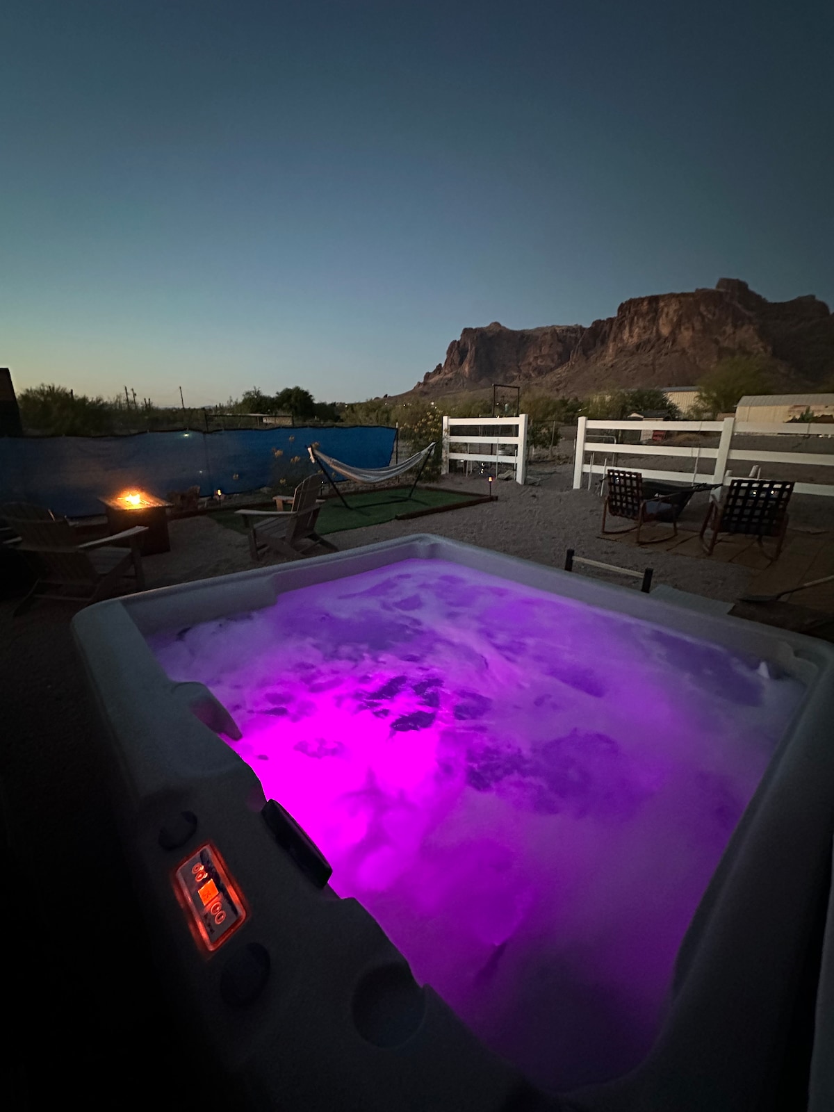 A private hot tub is illuminated with soft purple lights, surrounded by a tranquil outdoor space. A cozy campfire and lounge chairs complement the scene, with desert mountains visible in the background under a twilight sky.