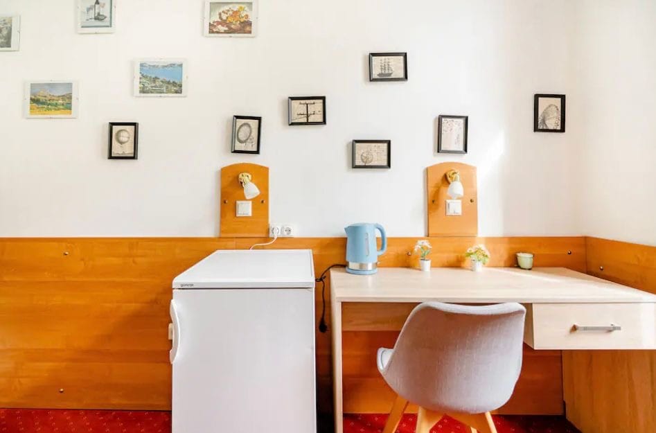 A bright room is presented with a small desk and a comfortable chair, accompanied by a compact refrigerator. Several framed pictures are arranged on the wall, providing a subtle decorative touch. A kettle rests on the desk, adding convenience for refreshments.